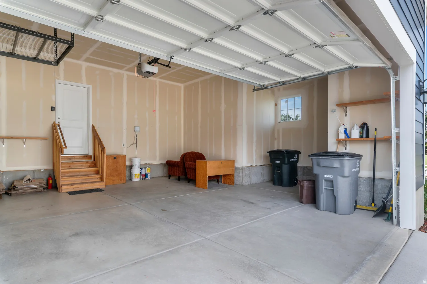 Concrete flooring, overhead storage rack, interior access door with wooden stairs, elevated wall shelves, and a window for natural light