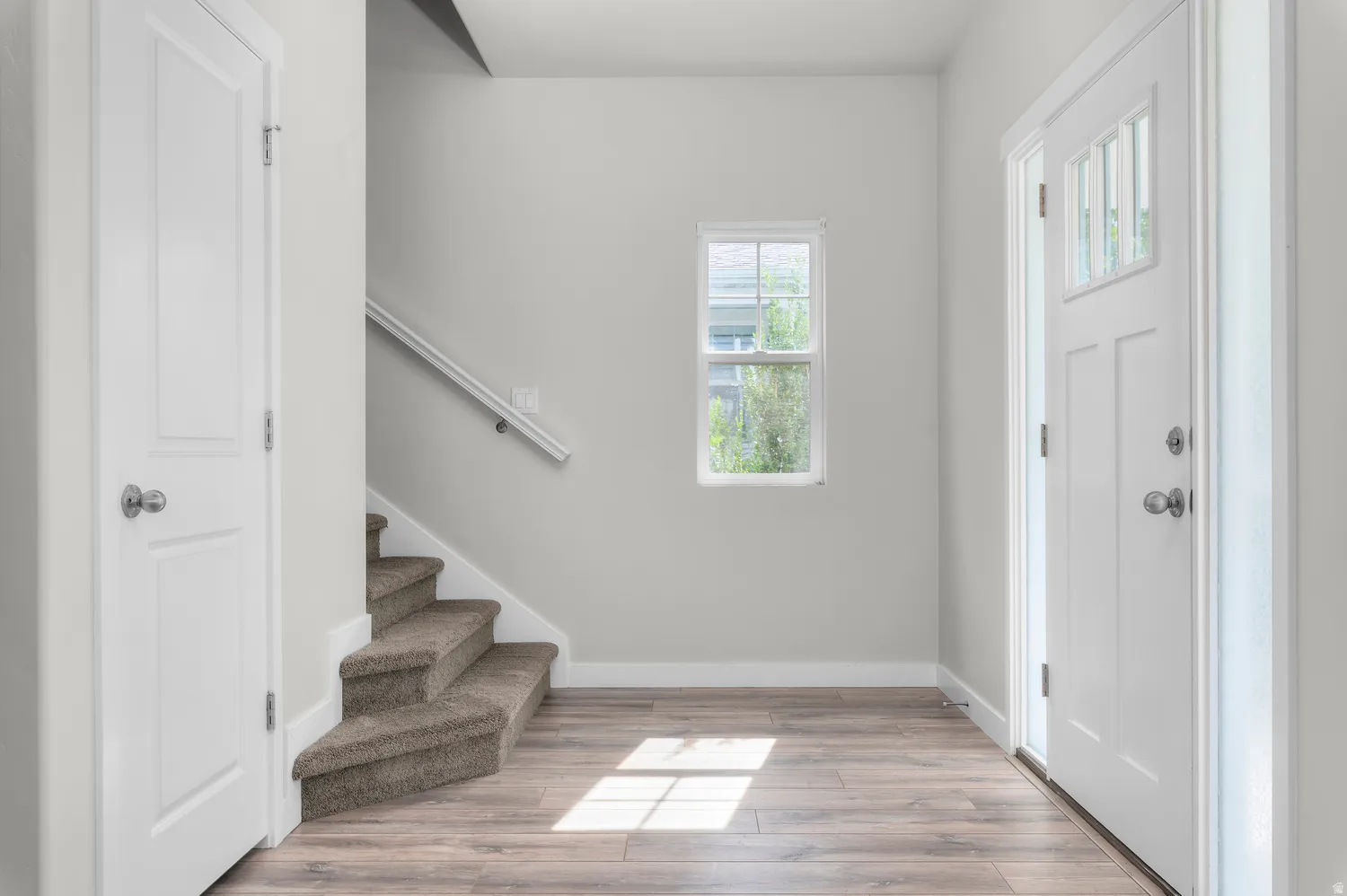 Entryway featuring wood-finish flooring, a contemporary front door with clear glass panels, a single window, and carpeted stairs with a white handrail