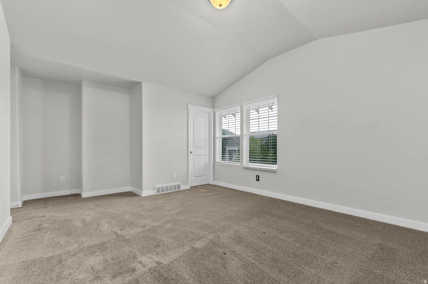 Neutral-toned carpeting, vaulted ceiling, dual windows with blinds, recessed wall features, and white trim throughout