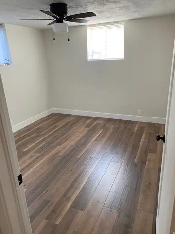Expansive room with wood-finish flooring, light-toned walls, a ceiling fan, and window