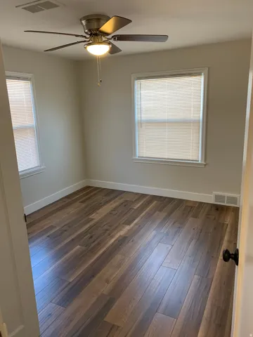 Wood-finish flooring throughout, two windows with blinds, a ceiling fan with integrated lighting, and white baseboards