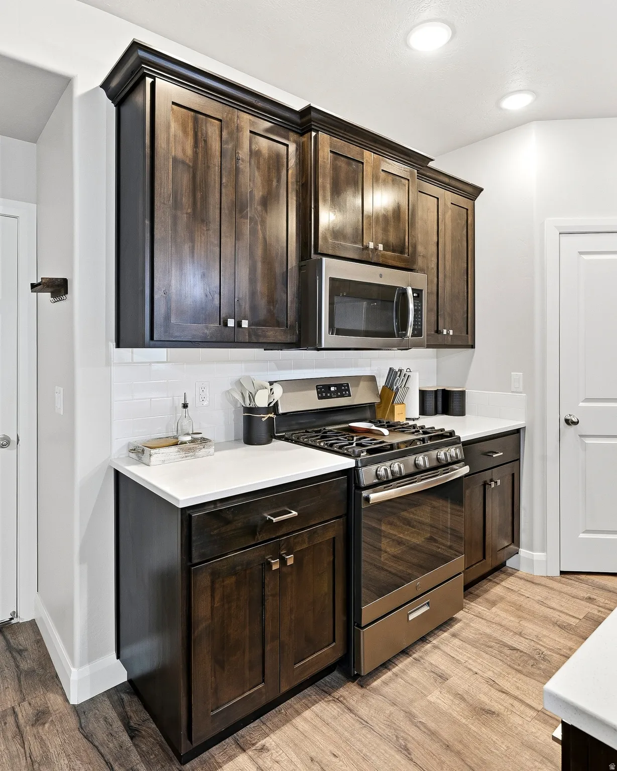 Dark wood-finish cabinetry with crown molding, stainless steel appliances, white subway tile backsplash, light-toned countertops, and wood-finish flooring