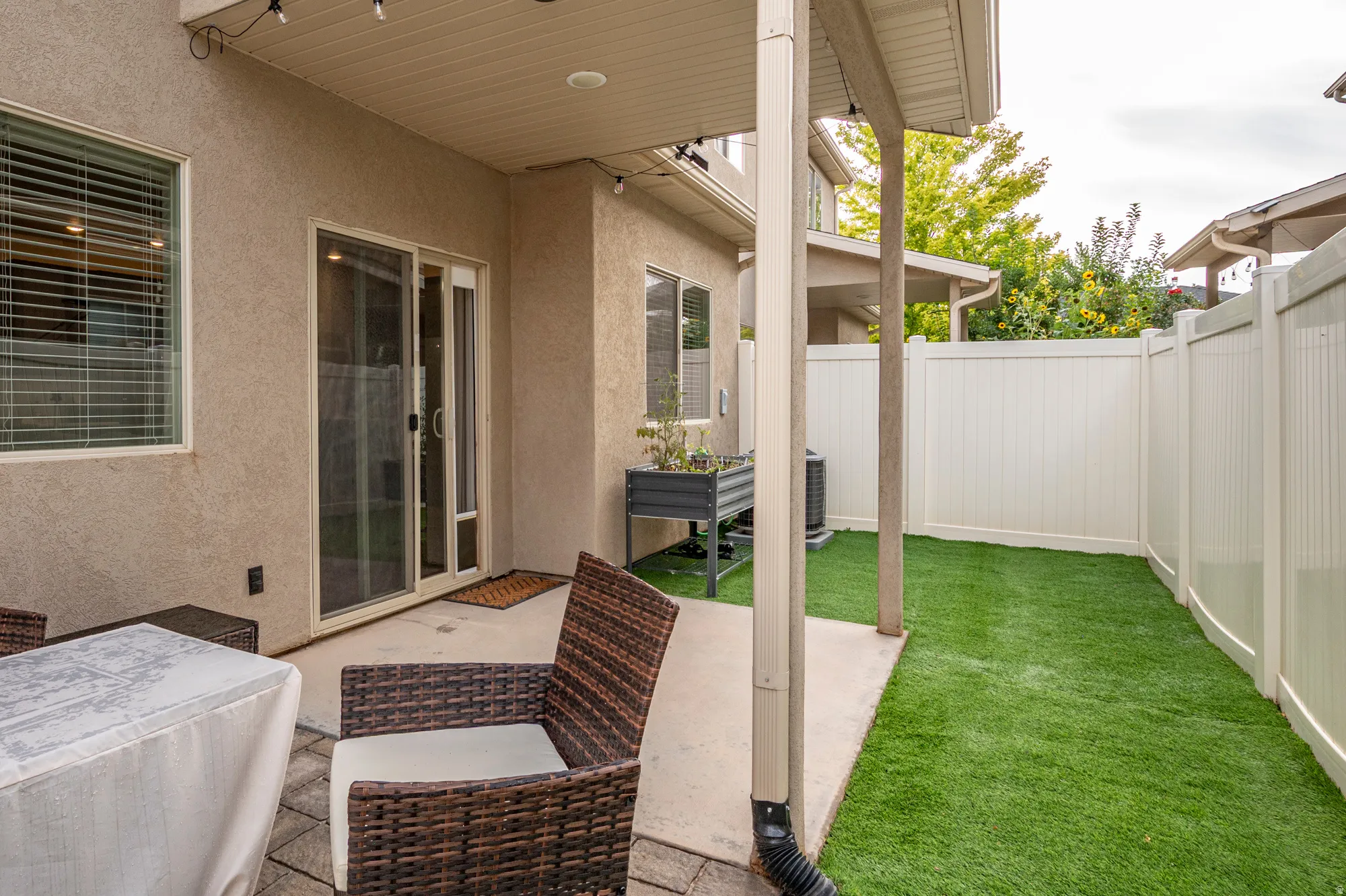 Covered patio area with concrete and paver flooring, sliding glass door access, enclosed by a privacy fence, and featuring artificial turf