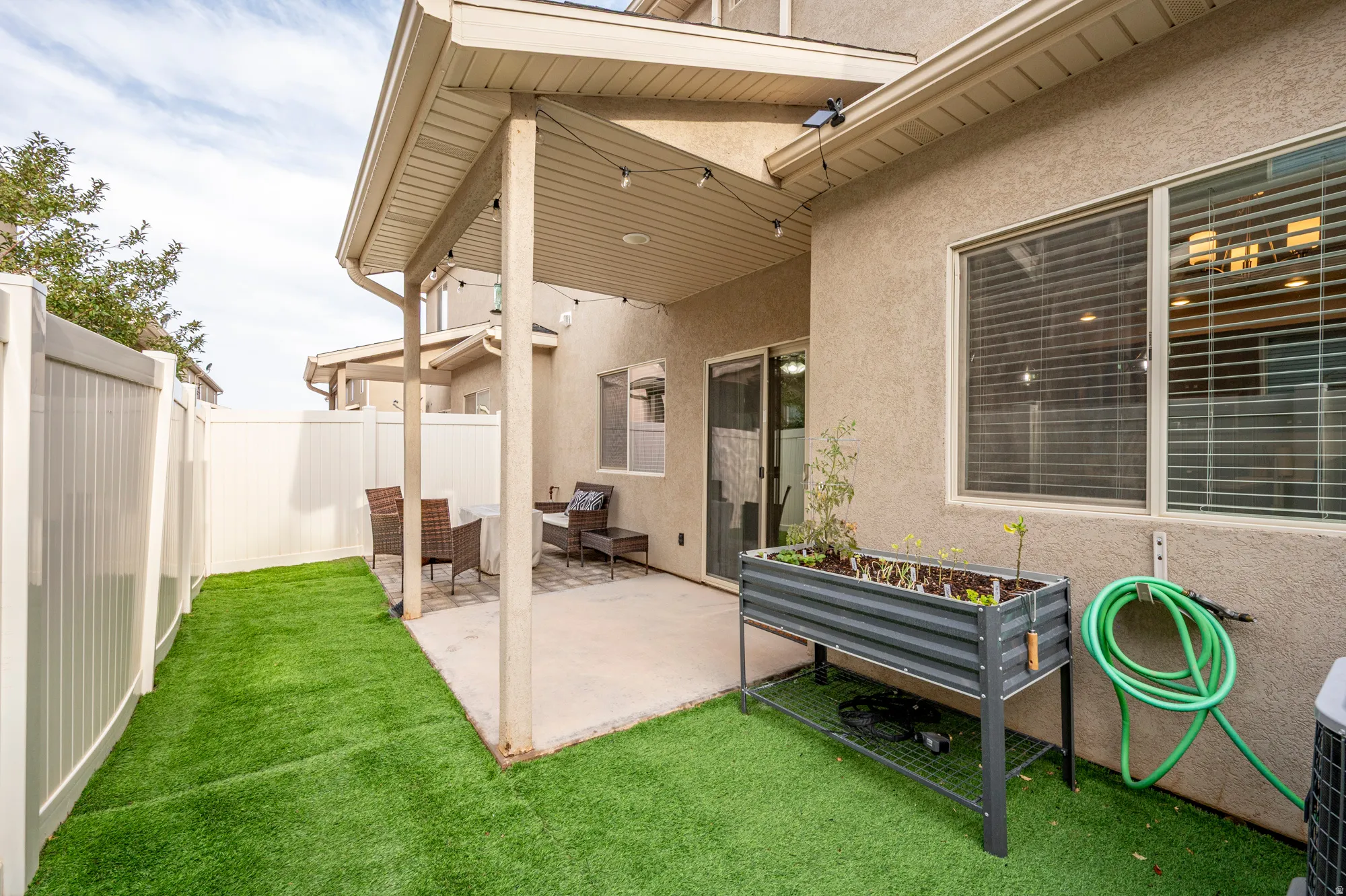 Covered patio with a concrete slab, artificial turf, white vinyl fencing, and an exterior wall with stucco finish