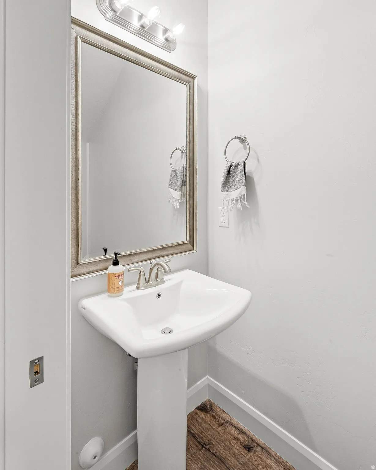 Pedestal sink with a brushed nickel faucet, framed mirror, wall-mounted light fixture, and wood-finish flooring