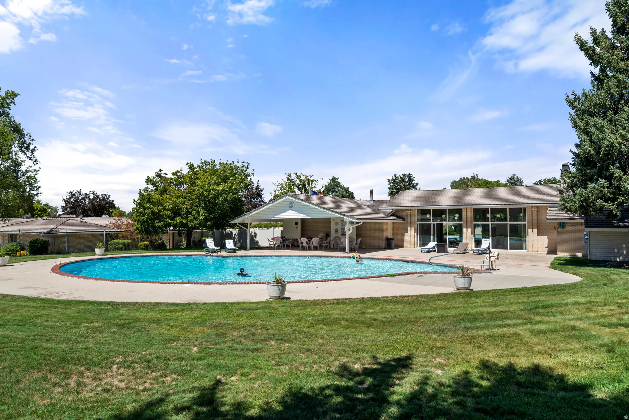 Expansive outdoor swimming pool with surrounding concrete decking, a covered patio area, and a large building featuring multiple glass-paned doors