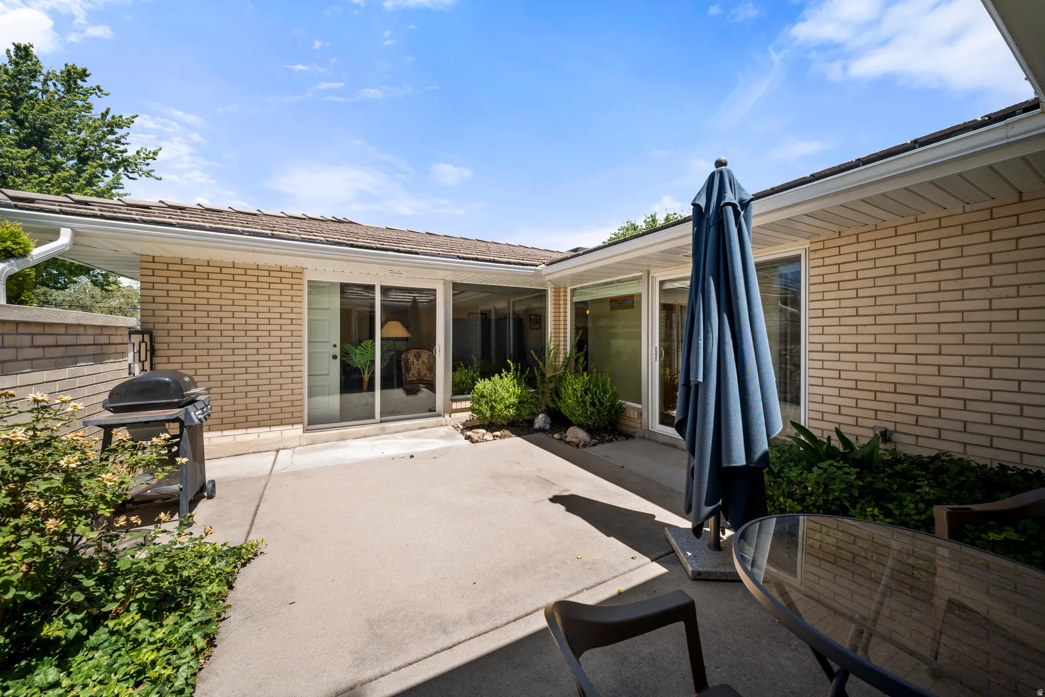 Paved patio area, light-colored brick exterior, sliding glass doors, roof overhang, integrated landscaping