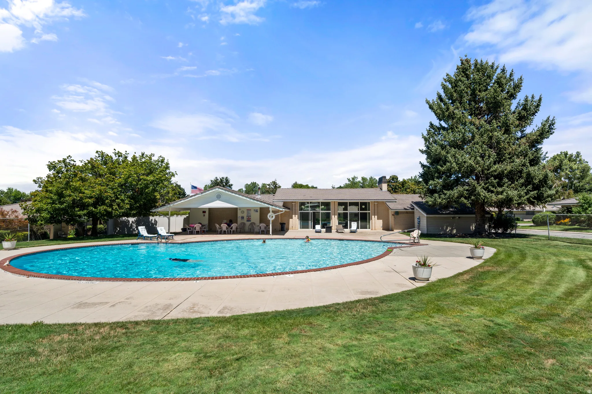 Community swimming pool with concrete surround, expansive lawn, mature landscaping, and an adjacent covered patio area