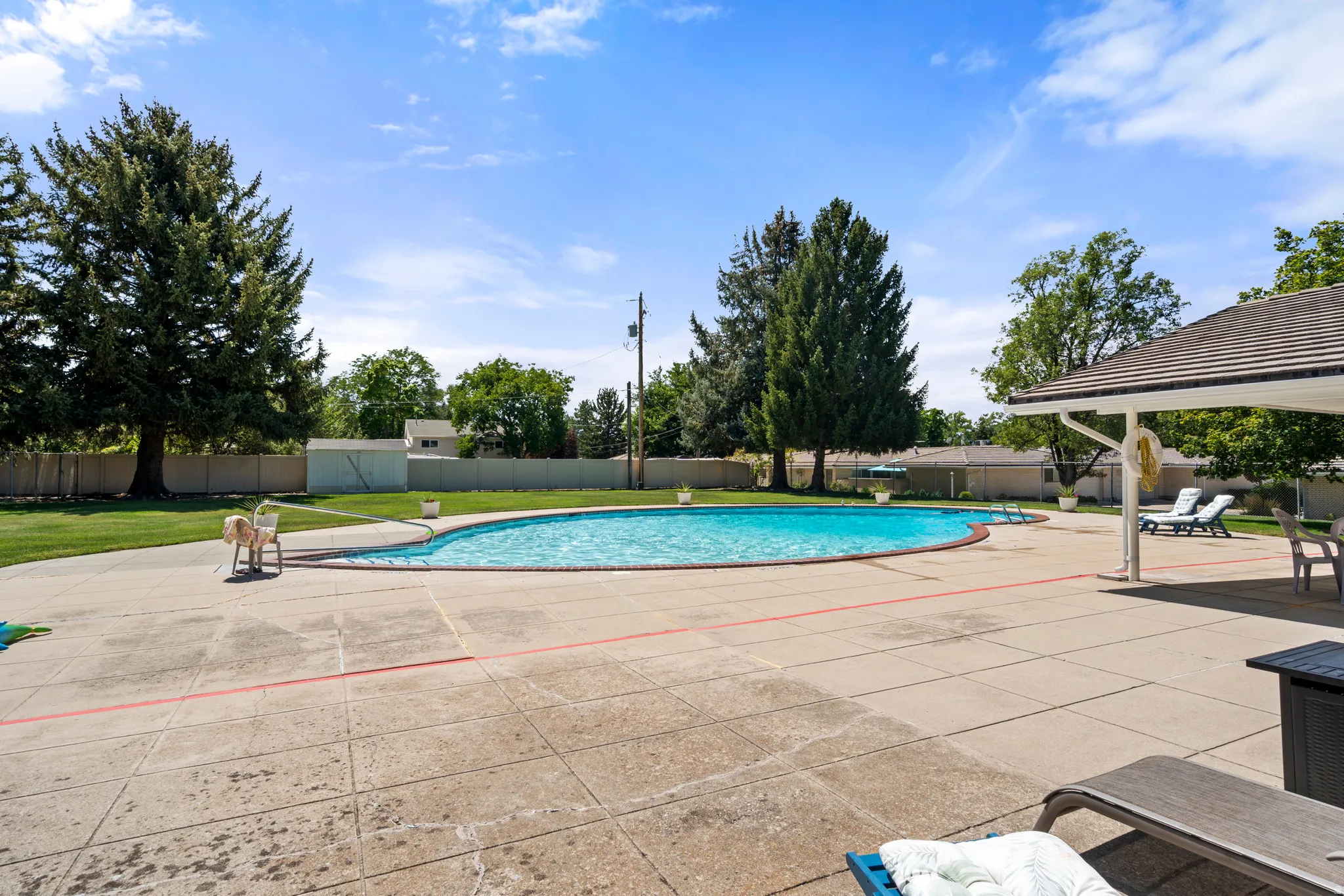 Large outdoor swimming pool with expansive concrete decking, mature perimeter trees, and a covered patio area