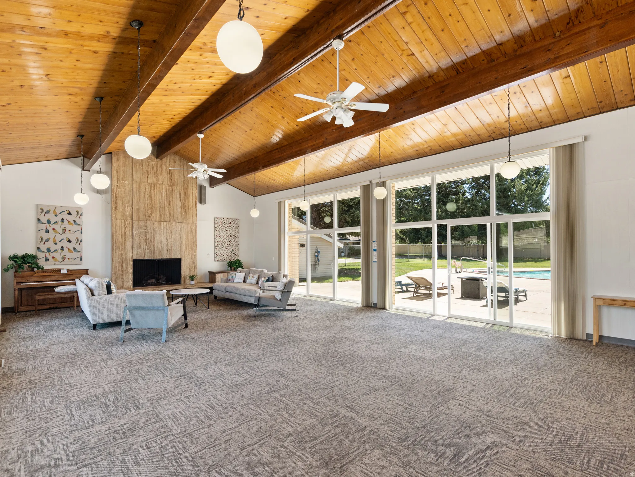 Vaulted wood-paneled ceiling with exposed beams, full-height fireplace with stone surround, wall of glass doors, integrated ceiling fans, and pendant lighting