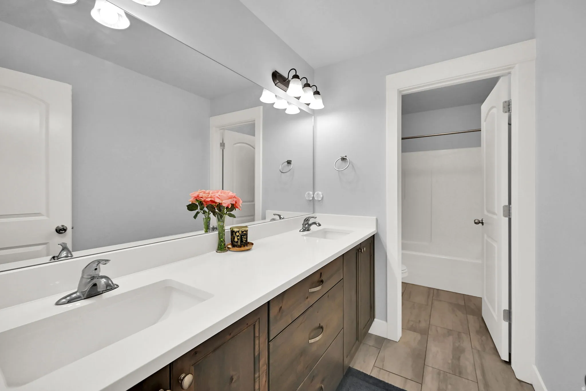 Dual vanity with integrated sinks, wood-finish cabinetry, a large wall-mounted mirror, and tile flooring