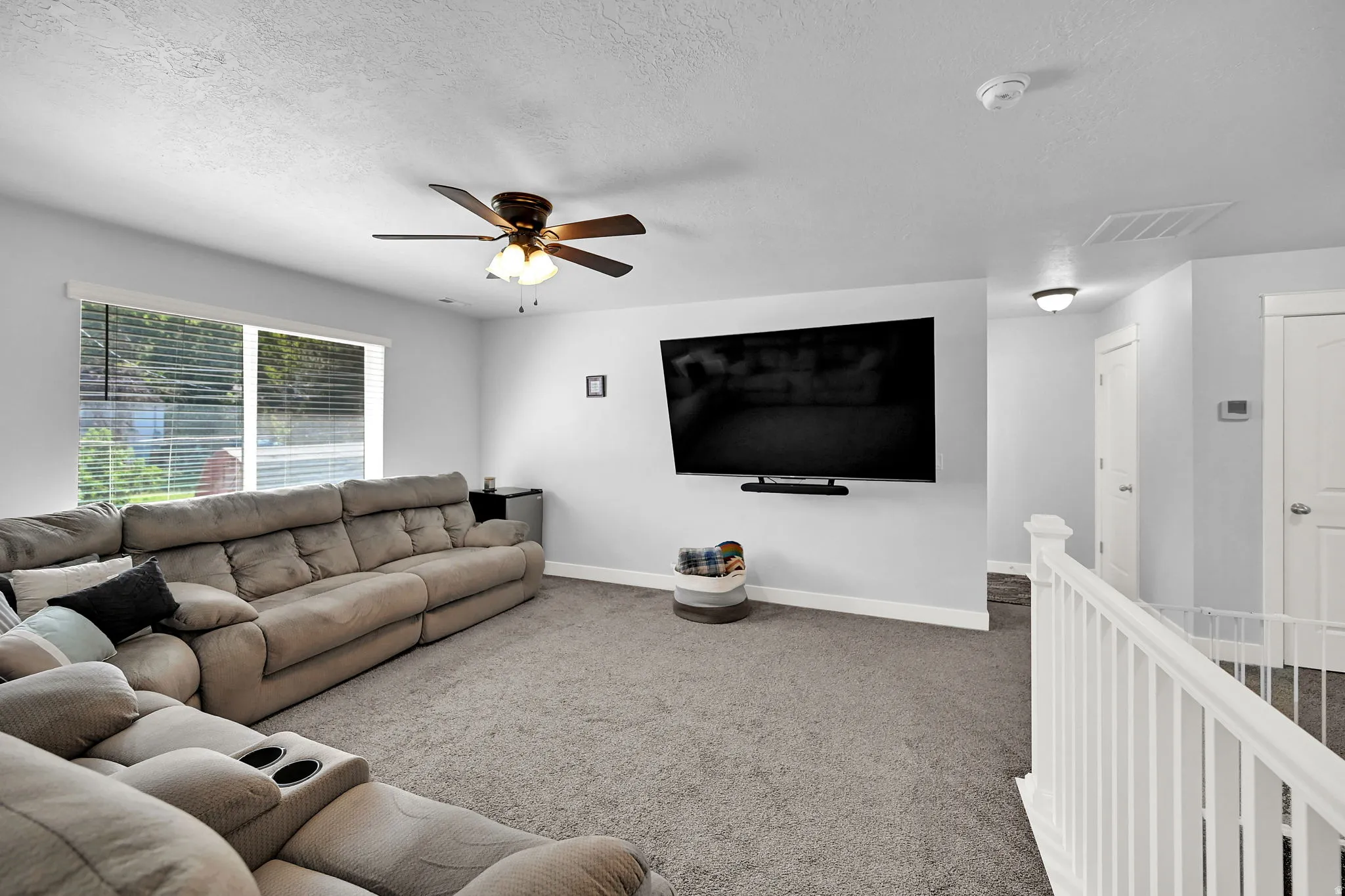 Carpeted living area with a ceiling fan, wall-mounted television, and a window with blinds
