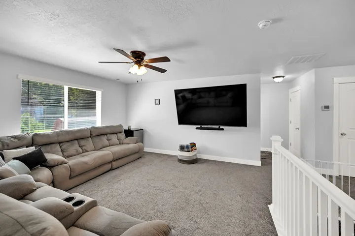 Carpeted living area with a ceiling fan, wall-mounted television, and a window with blinds