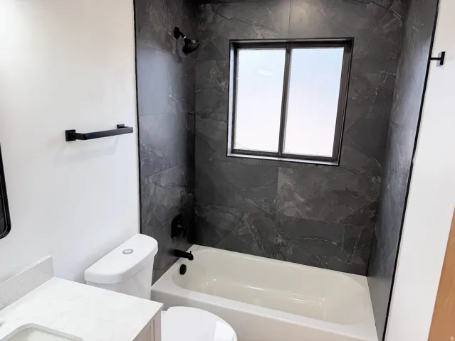 Bathtub with dark-toned tile surround, matte black shower fixtures, frosted window, and a modern toilet