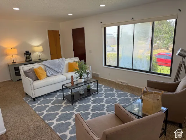 Recessed lighting illuminates this bright living space featuring a large picture window, light-toned walls, and neutral carpet flooring