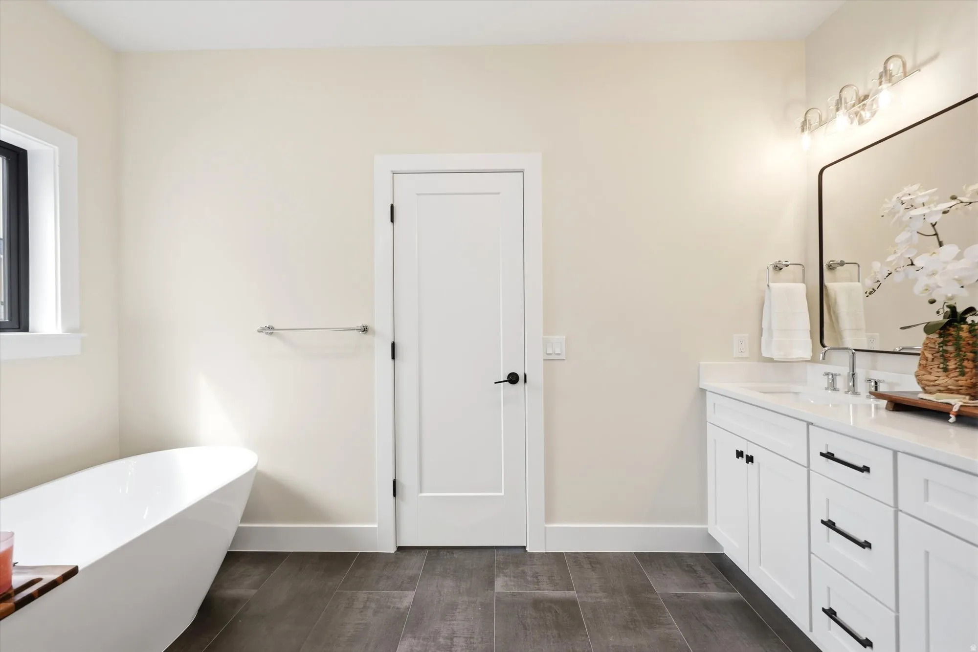 Freestanding soaking tub, white dual-sink vanity with dark hardware, framed rectangular mirror, and dark wood-finish flooring