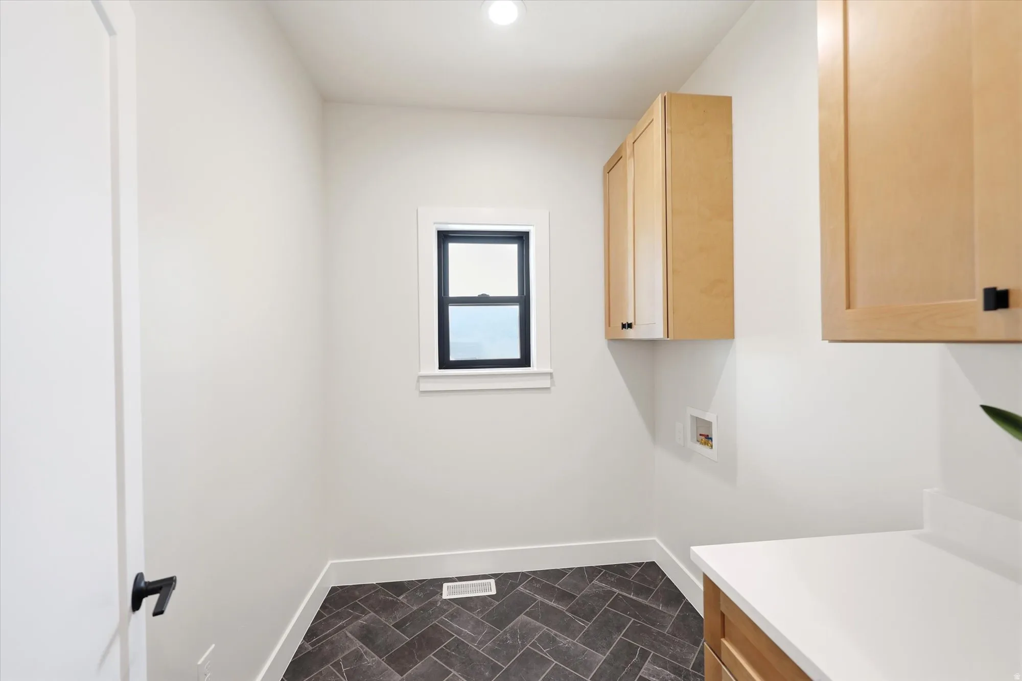 Recessed ceiling lighting, a window with black trim, wood-finish cabinetry, a white countertop, and dark herringbone-patterned floor tiles