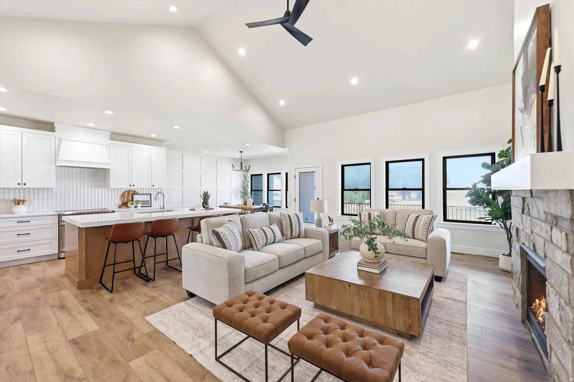 Vaulted ceiling with recessed lighting, wood-finish flooring, stone-clad fireplace, kitchen island with built-in sink, and white cabinetry