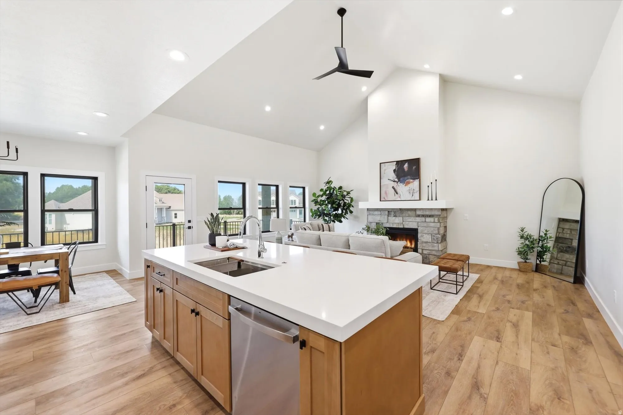 Vaulted ceiling with recessed lighting, wood-finish flooring, stone-faced fireplace with a mantel, kitchen island with a built-in sink and stainless steel dishwasher, and black-framed windows