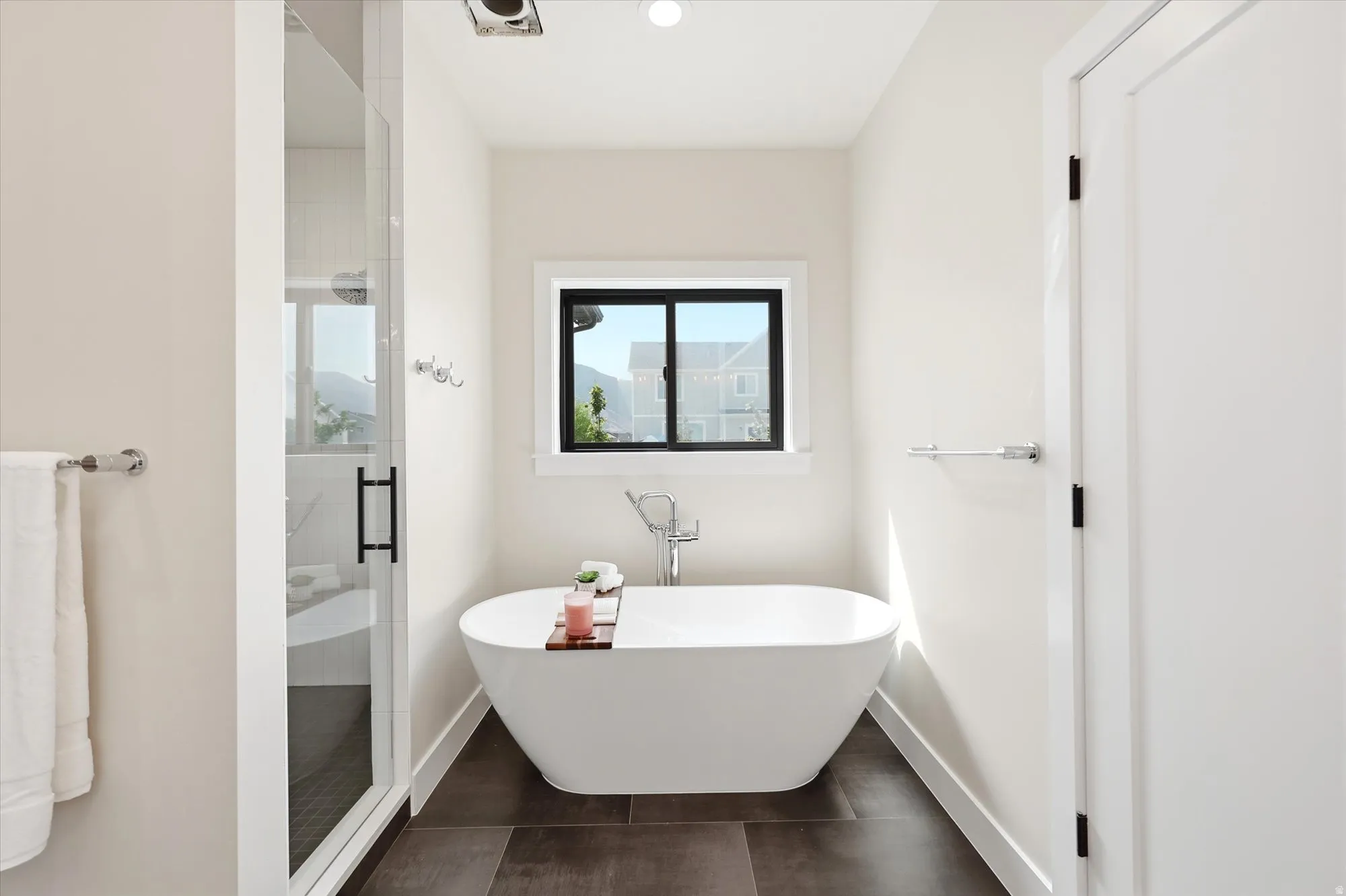 Freestanding soaking tub, glass-enclosed shower, dark tile flooring, and a large window with black trim