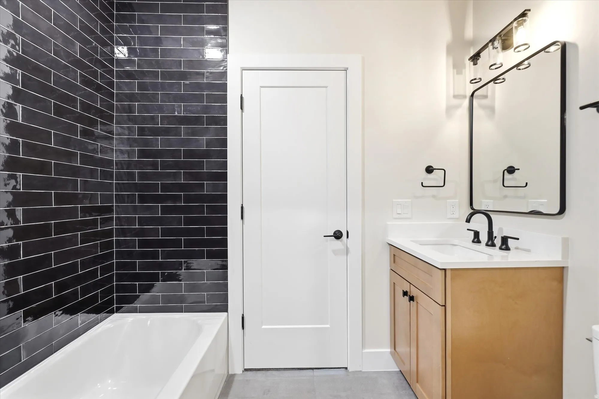 Subway tile shower surround, integrated bathtub, natural wood vanity, white countertop with integrated sink, and matte black fixtures