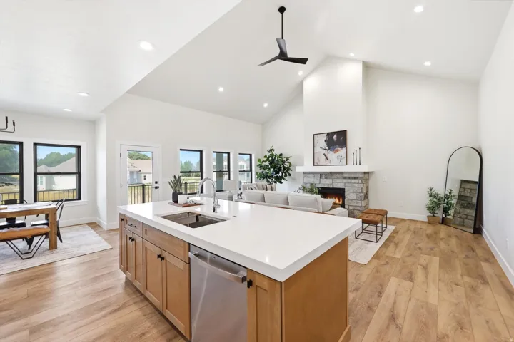 Vaulted ceiling with recessed lighting, wood-finish flooring, stone-faced fireplace with a mantel, kitchen island with a built-in sink and stainless steel dishwasher, and black-framed windows