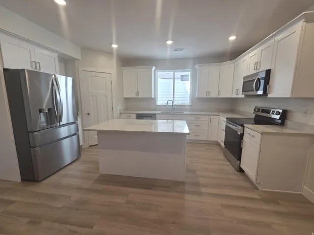 Contemporary kitchen featuring an integrated island, white shaker cabinetry, stainless steel appliances, light-toned countertops, and wood-finish flooring