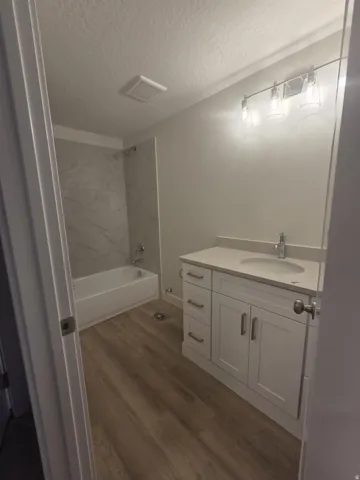 White vanity with integrated sink, wood-finish flooring, three-light vanity fixture, and a tiled shower/tub combination
