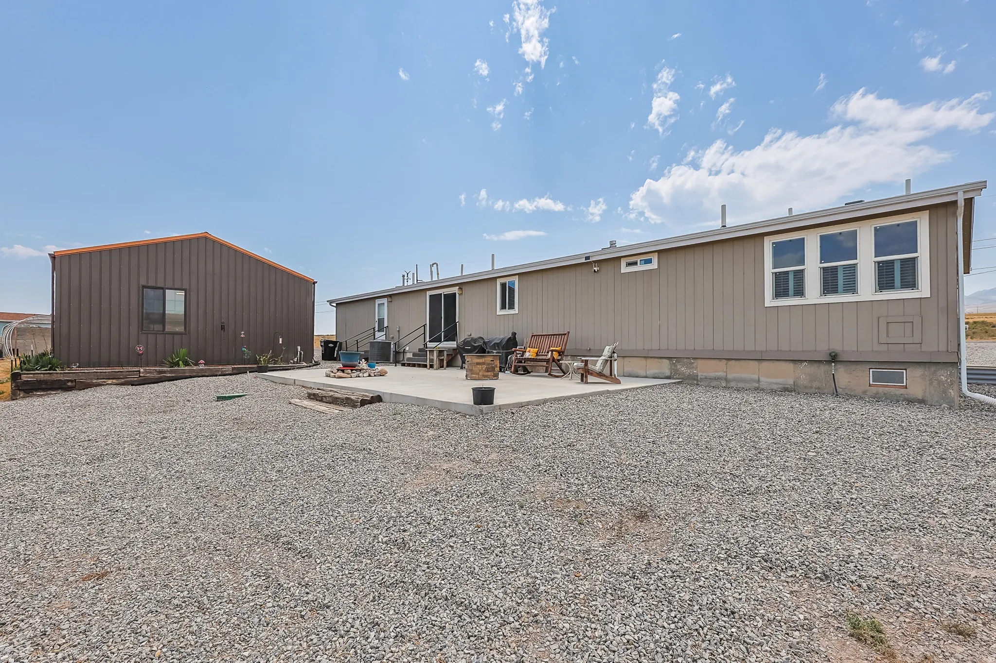 Expansive gravel lot with a concrete patio, a detached metal-sided building, and a tan exterior structure featuring multiple windows