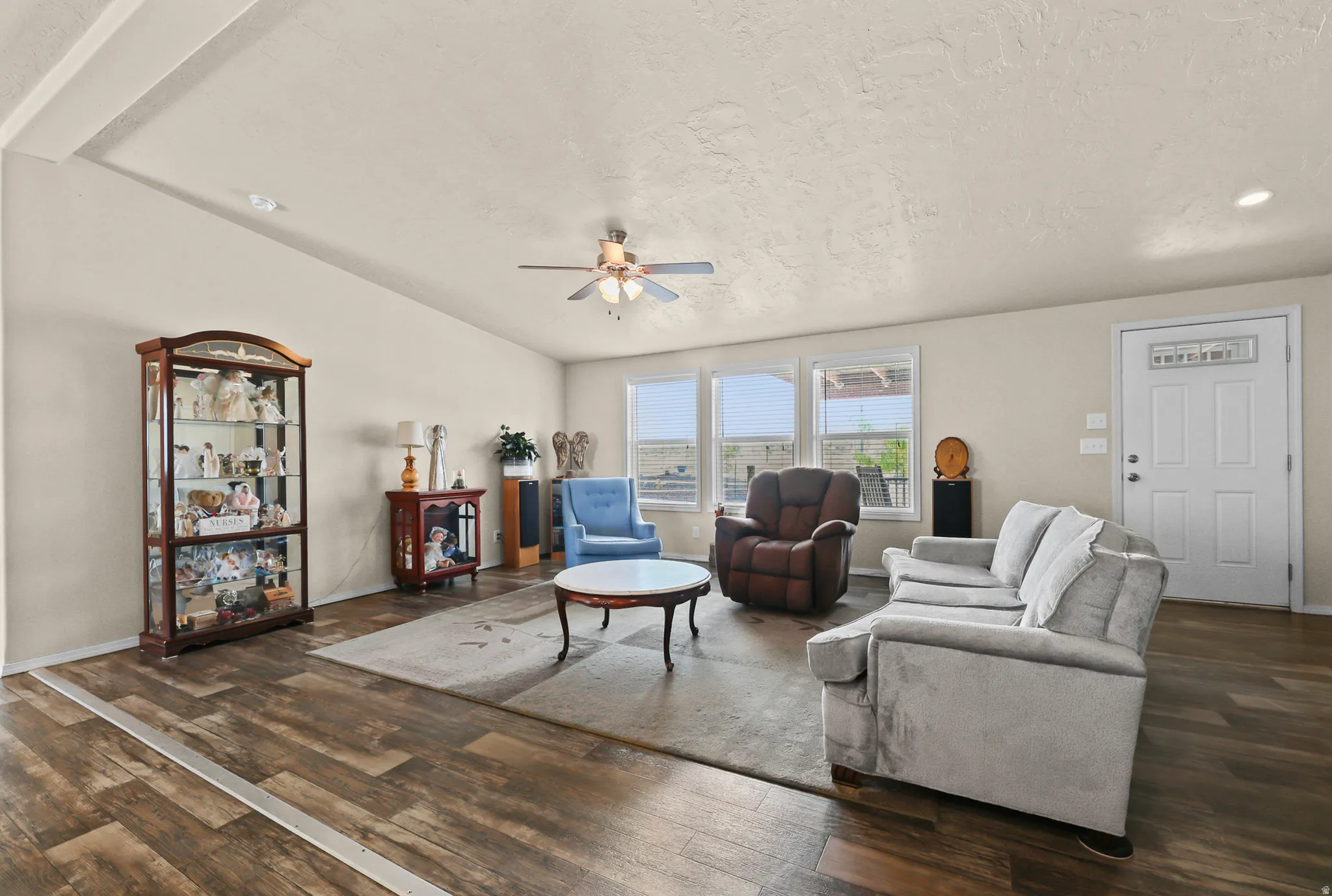 Spacious living area featuring wood-finish flooring, vaulted ceiling, ceiling fan, multiple windows, and a white paneled entry door with decorative glass