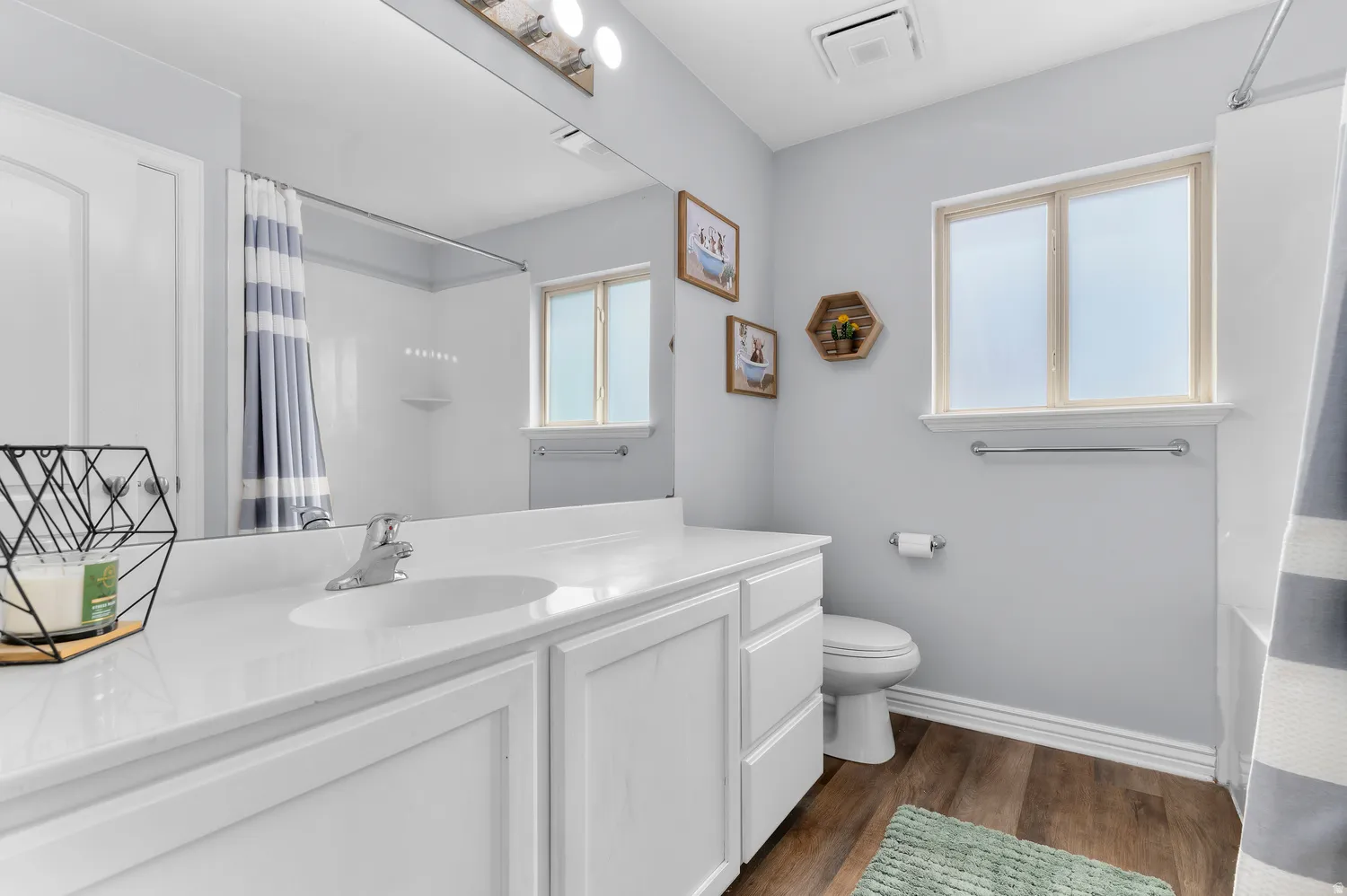Full-width vanity mirror, white shaker-style cabinetry, white solid surface countertop, wood-finish flooring, and a window with privacy glass