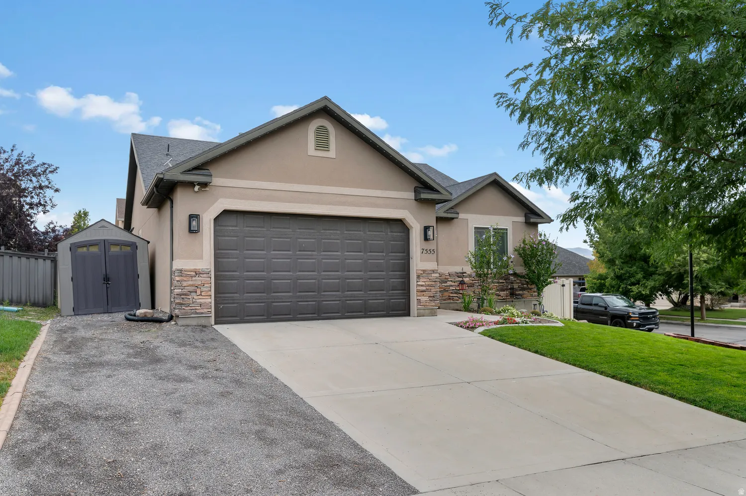 Stucco exterior with stone accents, a two-car garage, a concrete driveway, a separate gravel parking area, and a storage shed