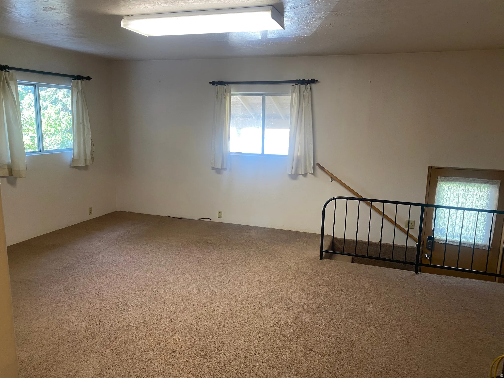 Wall-to-wall carpeting, two windows with curtain rods, fluorescent overhead lighting, and a wooden handrail with a metal railing
