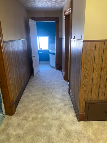 Carpeted hallway with wood-finish wainscoting and trim, leading to a room with a window and white cabinetry