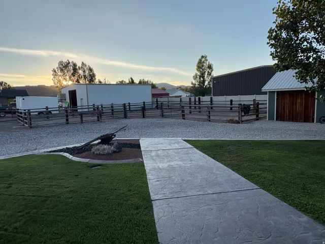 Stamped concrete pathway leading to a gravel area, featuring wood fencing, a metal-clad outbuilding, and a smaller shed with wood doors