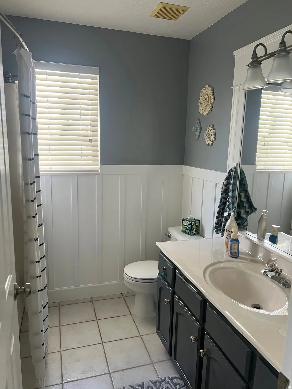 White board and batten wall paneling, window with horizontal blinds, tiled flooring, vanity with dark cabinetry, and a countertop with integrated sink