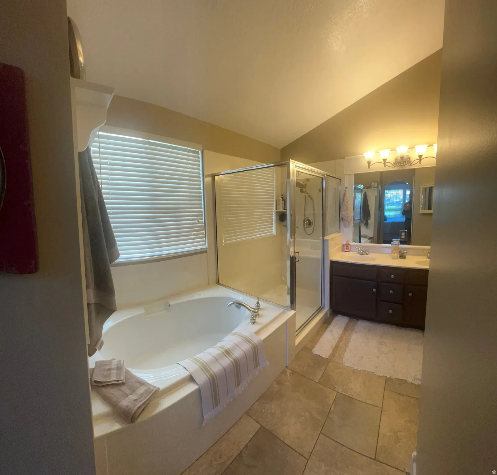 Soaking tub with a built-in shelf, glass-enclosed shower, dual-sink vanity with a solid surface countertop, and large format tile flooring