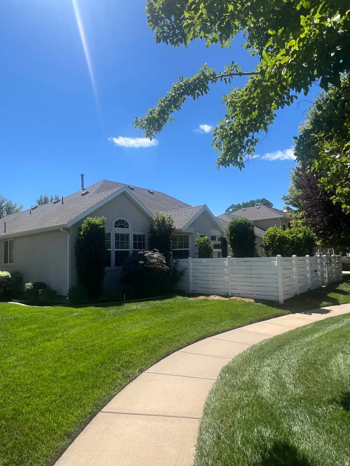Paved walkway bordering a manicured lawn, white horizontal slat fence, and mature trees