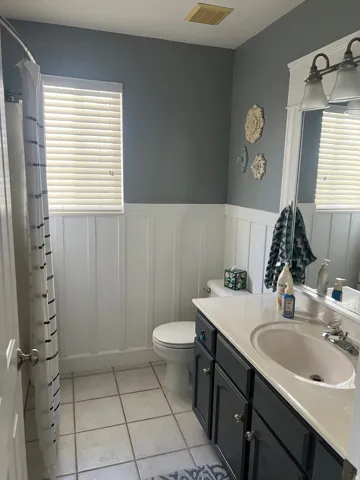 White board and batten wall paneling, window with horizontal blinds, tiled flooring, vanity with dark cabinetry, and a countertop with integrated sink