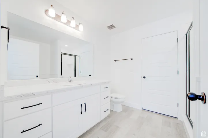 White double vanity with a single sink and black hardware, large frameless mirror, light wood-finish flooring, and a glass-enclosed shower