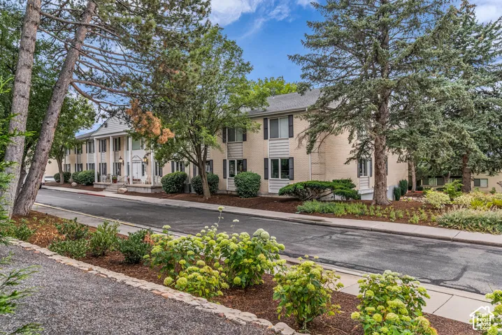 Two-story residential building with light-colored brick exterior, dark window shutters, established trees, and landscaped grounds featuring varied plantings