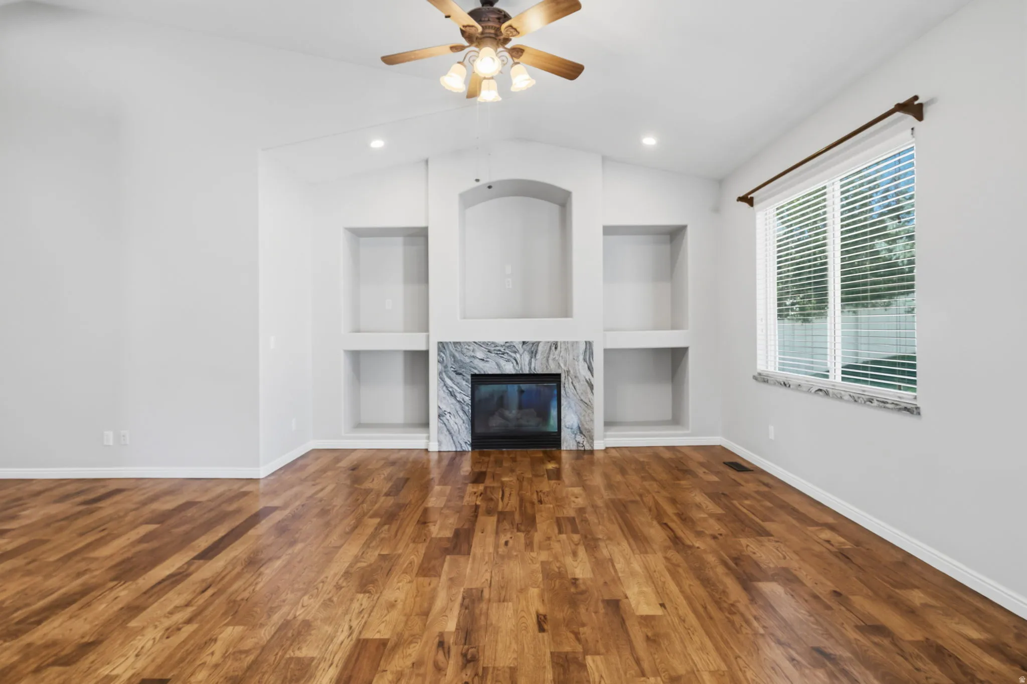 Vaulted ceiling with a ceiling fan, fireplace with a stone surround, built-in shelving, wood-finish flooring, and a large window with blinds
