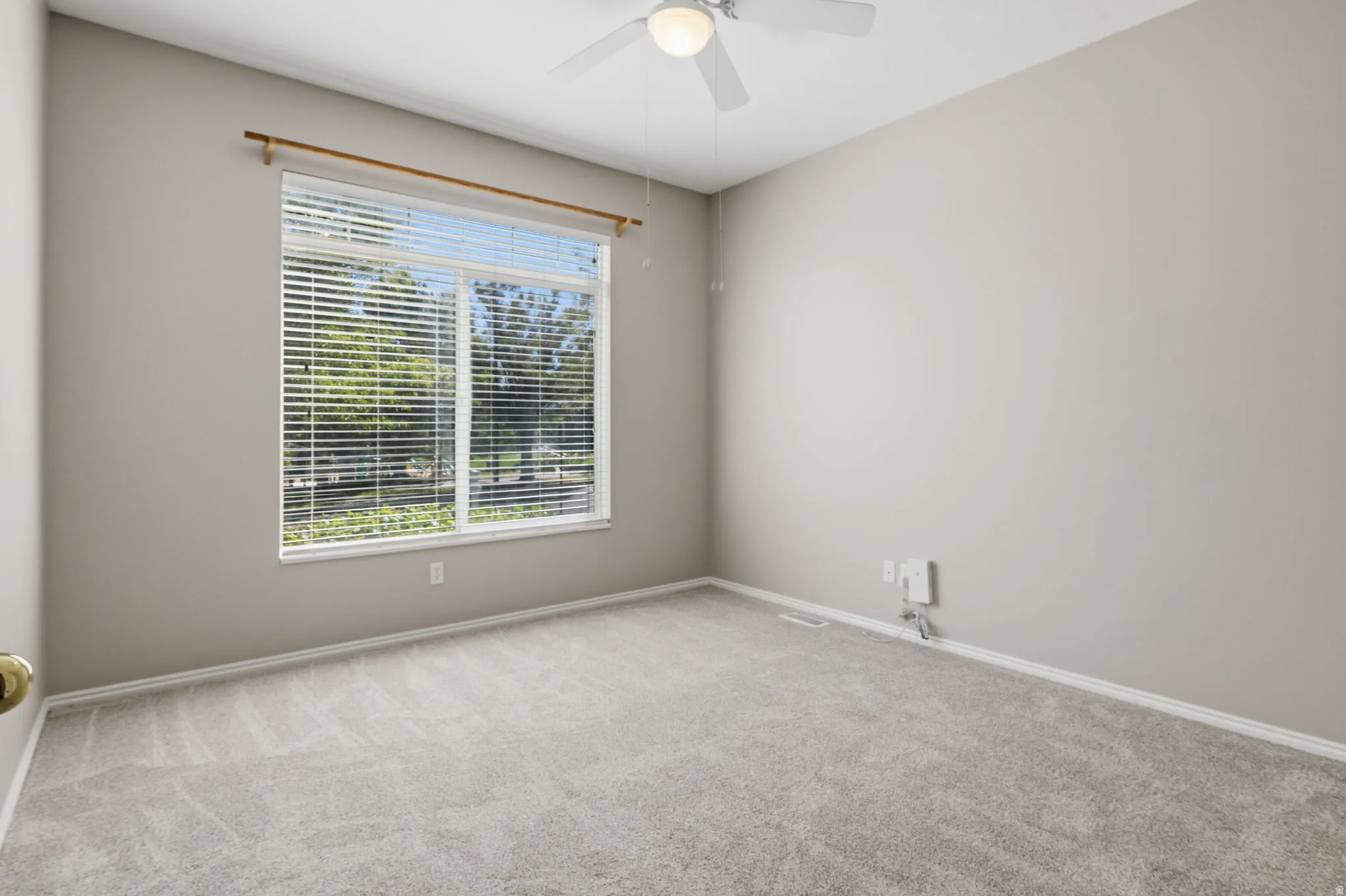 Neutral tone wall paint, carpeted flooring, large window with horizontal blinds, and a ceiling fan with integrated lighting