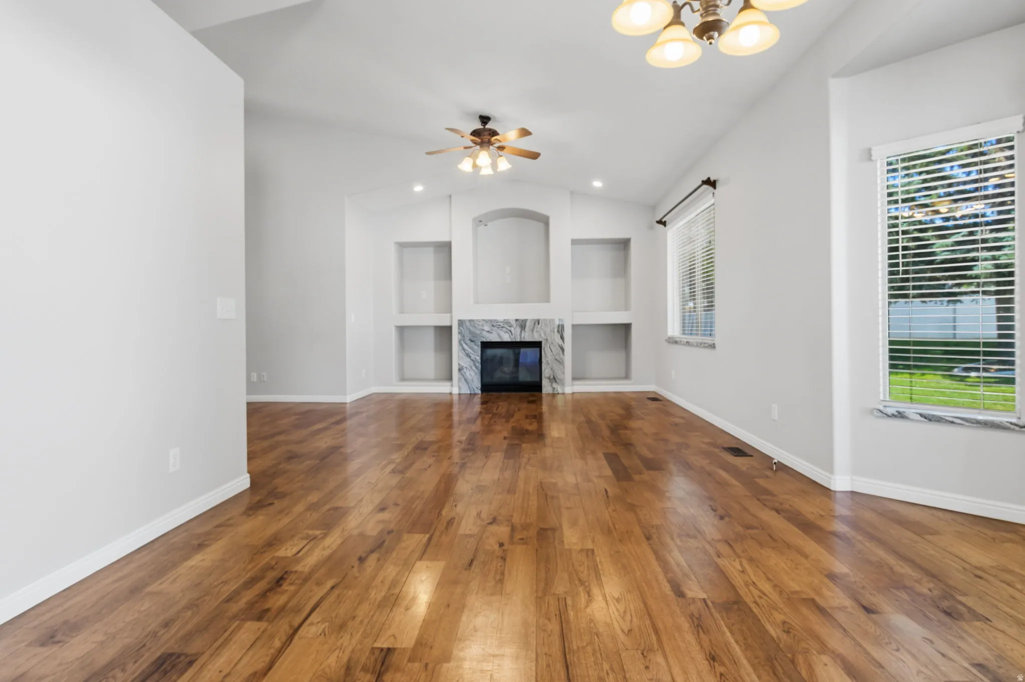 Vaulted ceiling with a ceiling fan, wood-finish flooring, built-in shelving, fireplace with a marble-look surround, and window with a marble-look sill