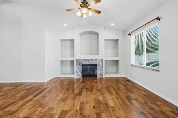 Vaulted ceiling with a ceiling fan, fireplace with a stone surround, built-in shelving, wood-finish flooring, and a large window with blinds