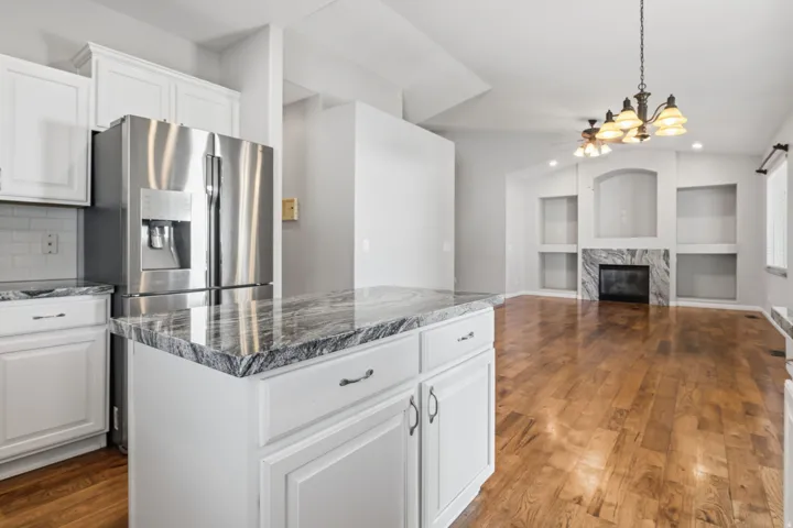 Stainless steel refrigerator, white cabinetry, island with stone-finish countertop, wood-finish flooring, and a fireplace with stone-finish surround