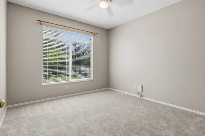 Neutral tone wall paint, carpeted flooring, large window with horizontal blinds, and a ceiling fan with integrated lighting