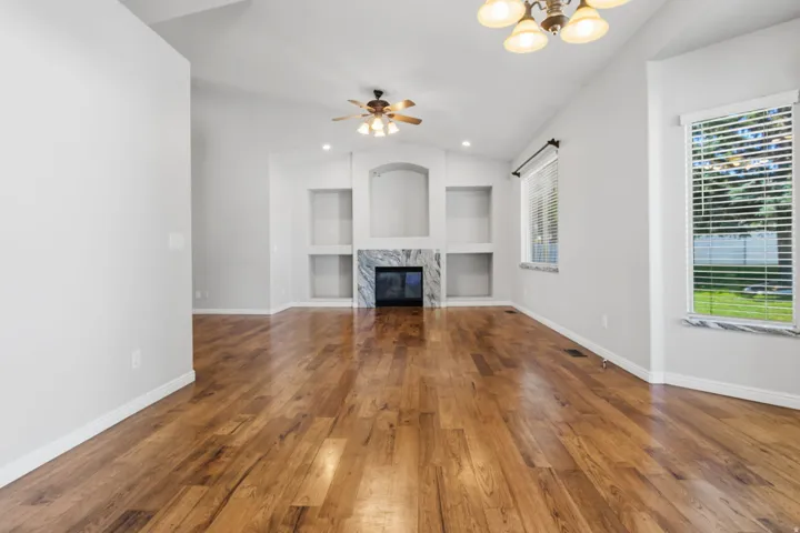 Vaulted ceiling with a ceiling fan, wood-finish flooring, built-in shelving, fireplace with a marble-look surround, and window with a marble-look sill