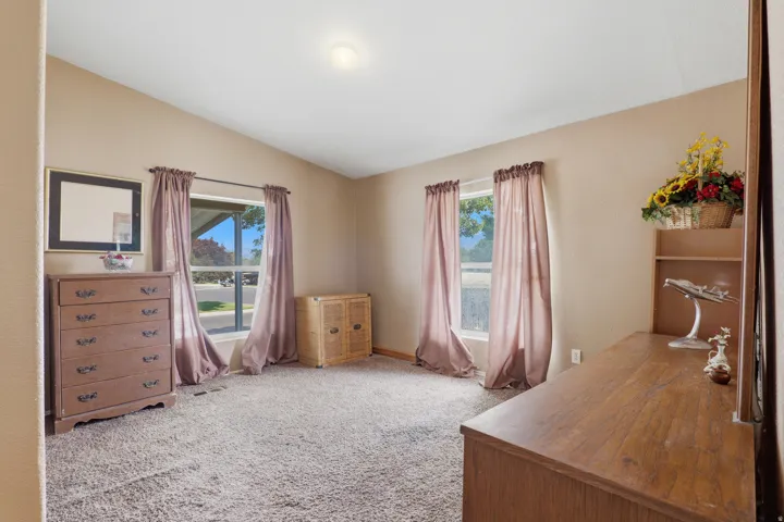 Spacious bedroom featuring wood-finish flooring, plush carpeted seating area, multiple windows with mauve drapery, wood dresser with mirror, and wood desk with shelving