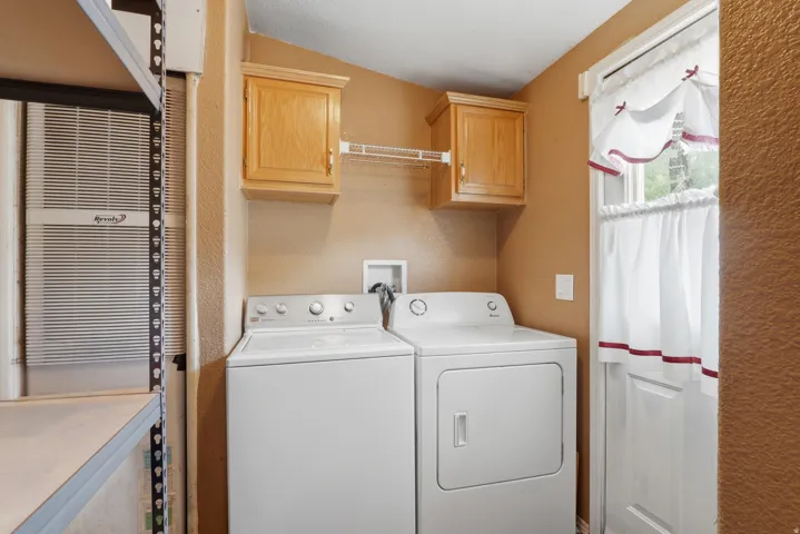 Dedicated laundry room featuring white front-load washer and electric dryer, wood-finish cabinetry with overhead storage, ceramic tile countertop, and natural light from a side window with decorative valance